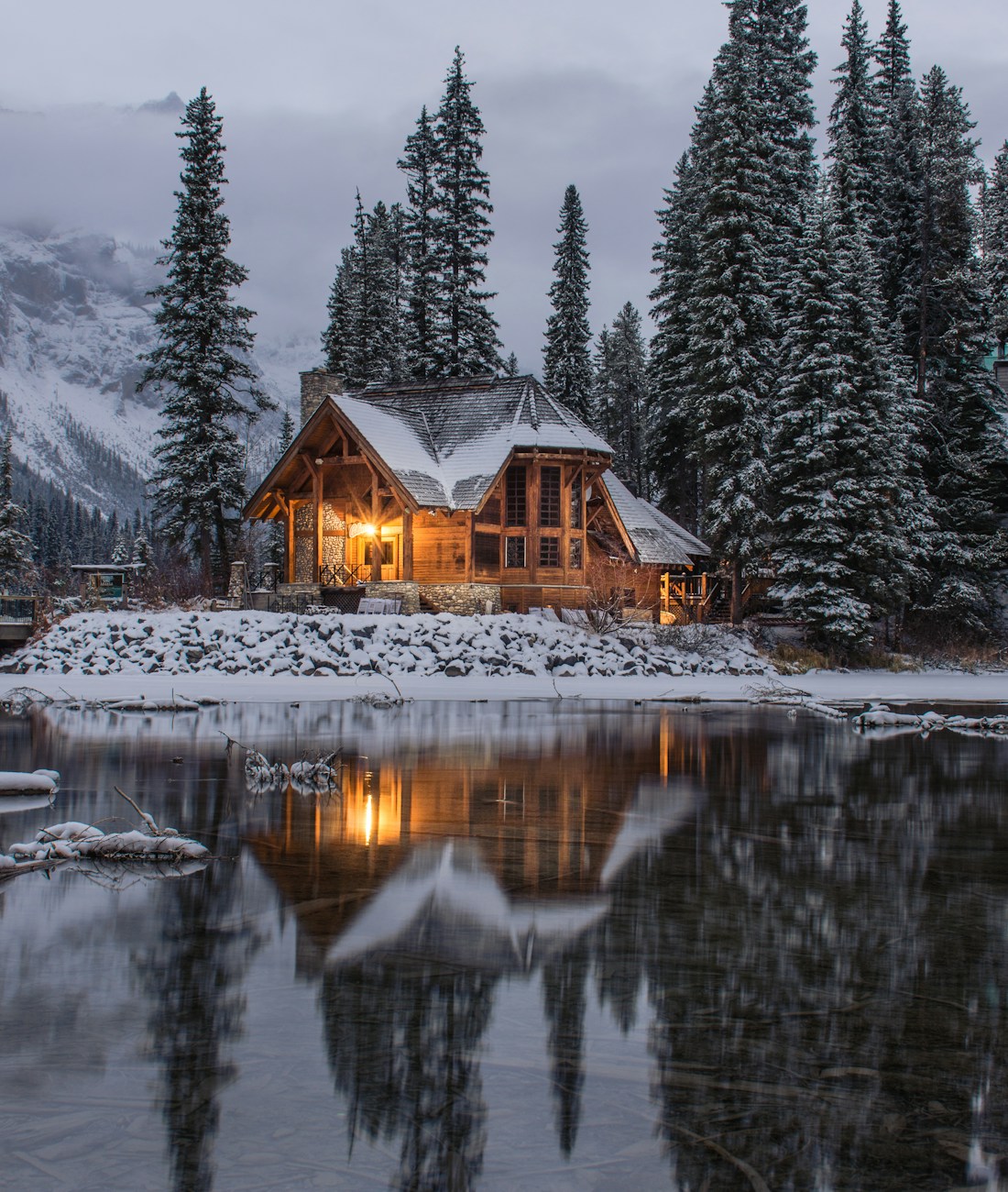 A timber mountain lodge glowing at dusk beside a still lake, snow on the roof