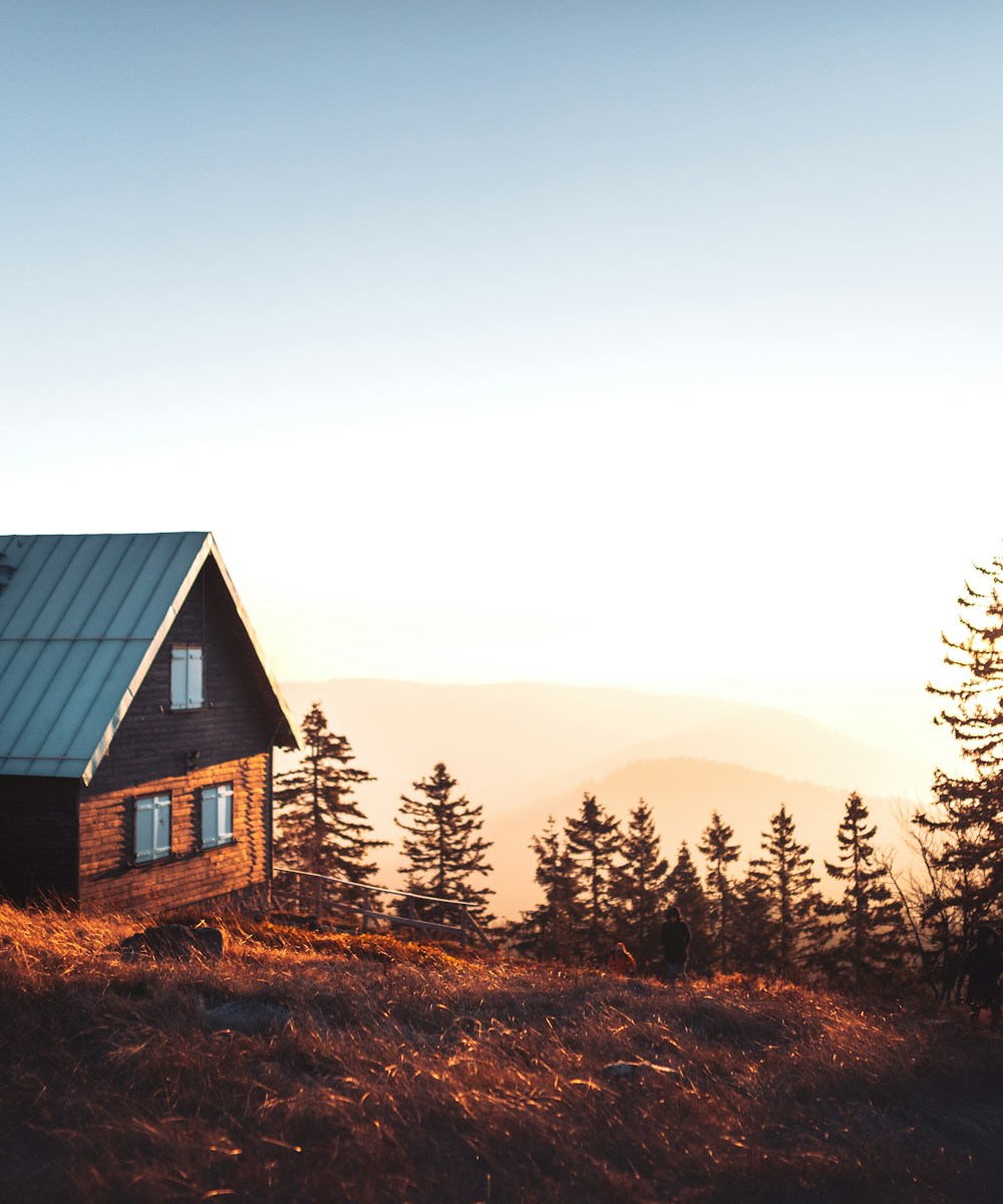 A dark-timber cabin on a hillside with the sun setting behind the pines