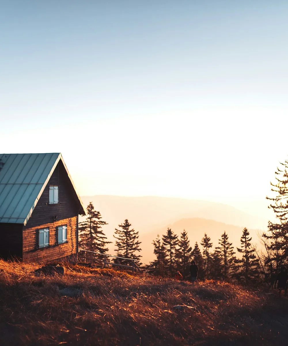A dark-timber cabin on a hillside with the sun setting behind the pines