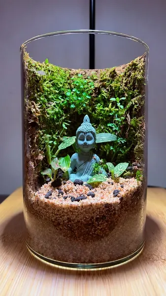 Glass cylinder terrarium with a Buddha sitting in a moss hollow above river sand